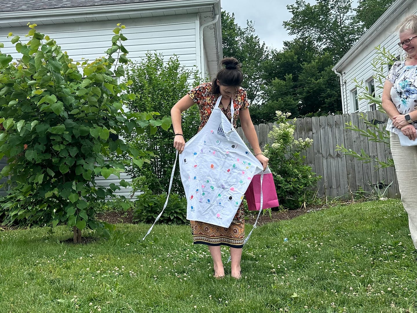 Lucy Apron 1 Two women in a backyard garden near a white house with a blue door and a Hilltop Montessori sign.